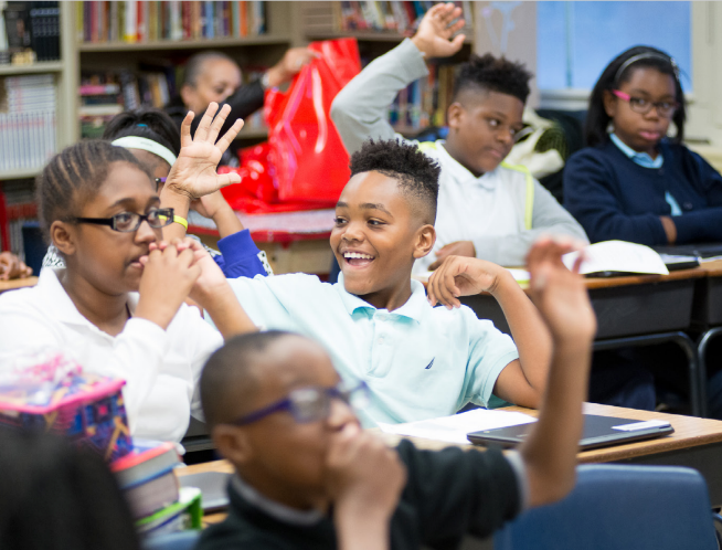 zoomed in landscape image of kids in class with one kid smiling and raising his hand