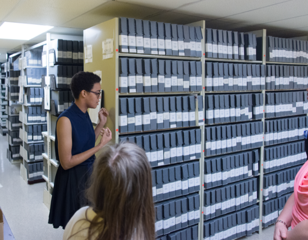 Woman standing in front of archive shelves talking to at least 2 people